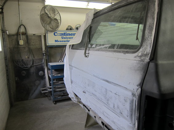 Side view of a vehicle body being prepped for paint in a workshop, with a fan and shelves in the background