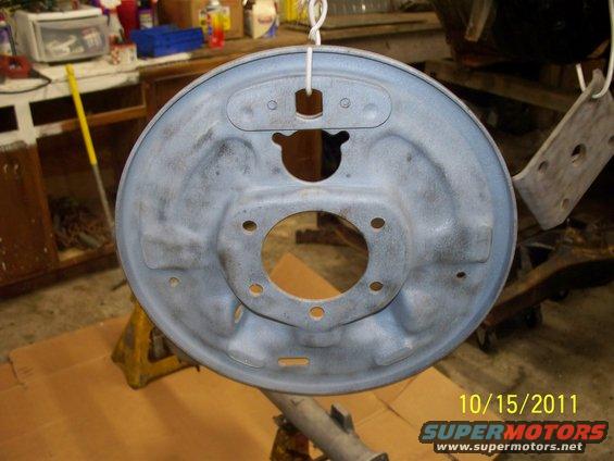 Gray brake drum backing plate hanging by a wire, showing bolt holes and a central opening