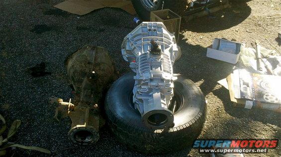 Two vehicle transmissions, one silver and one older brown, sit on the ground next to a tire. Boxes and other parts are visible in the background