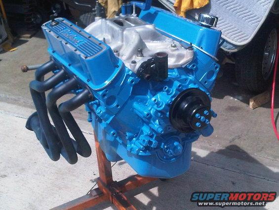 Blue V8 engine on an orange stand, with black headers and intake manifold. Supermotors.net logo in corner