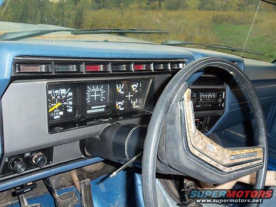 Dashboard interior of a blue vehicle with a wood-grain trim steering wheel and analog gauges. A screwdriver is in the ignition