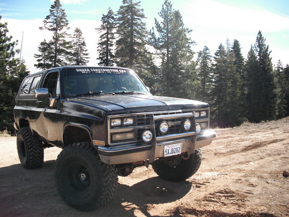 Lifted black 1985 Chevrolet K5 Blazer with off-road tires parked on a dirt road, trees in background