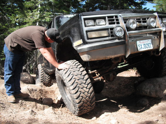 Man checks large off-road tire on lifted black truck with aftermarket bumper and lights. California plate 5XJB282
