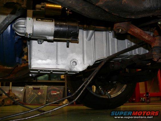 Car's starter motor bolted to the oil pan, with suspension parts and a wheel visible