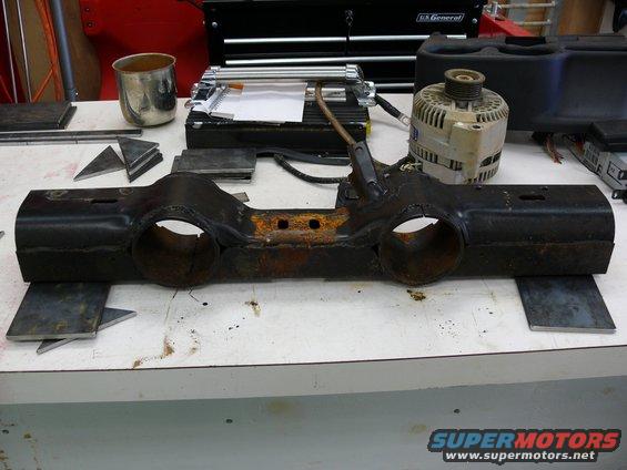 Rusty metal automotive part with two large circular openings and mounting holes, resting on a workbench with tools