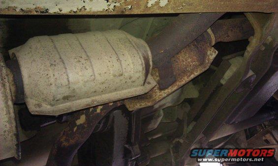 Underside view of a vehicle's exhaust system, showing the catalytic converter and pipes. Some rust is visible on the components