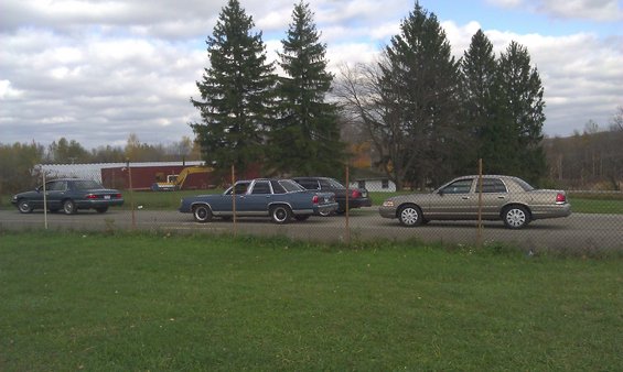 Four sedans parked on a road. The closest one is a gold Ford Crown Victoria. Behind it is a blue sedan, then a dark sedan, and a dark green sedan further back