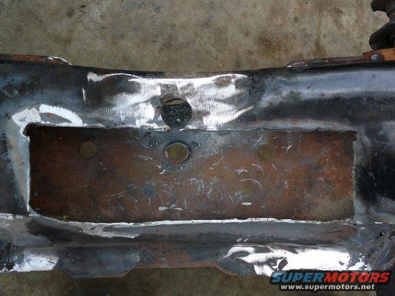 Close-up of a rusty, welded car frame section with visible circular indentations and a rectangular cutout