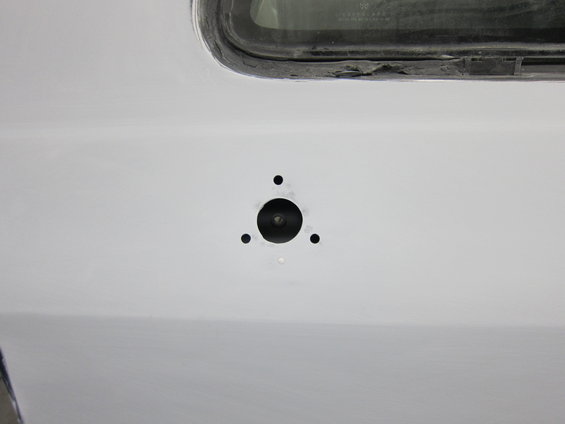 Close-up of a white vehicle body panel with a circular hole and three smaller surrounding holes, near a window seal