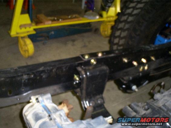 Close-up of a vehicle's frame with suspension parts and a large tire visible. A yellow engine hoist is in the background