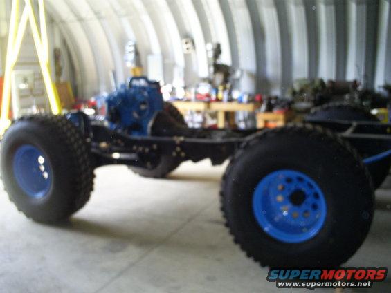 Vehicle chassis with a blue engine and large off-road tires with blue rims, viewed from the side in a garage