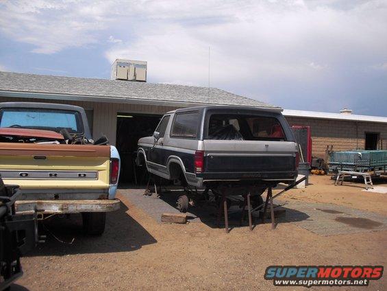 Two-tone Ford Bronco on jack stands, rear view. A Ford pickup truck is parked to the left