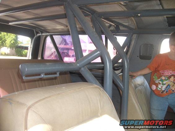 Interior view of a vehicle with a grey roll cage installed over tan seats. A child in an orange shirt stands to the right