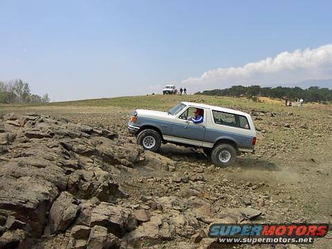 Two-tone blue and white Ford Bronco climbing rocky terrain. Another white SUV is visible in the distance