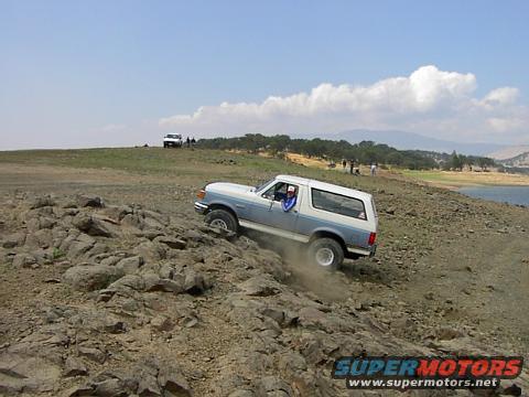 Blue and white 1988 Ford Bronco driving on rocky terrain near water