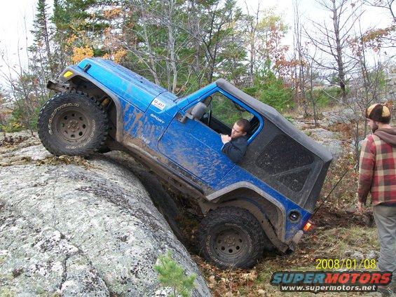 Blue Jeep Wrangler climbing over a large rock on a trail, driver inside, another person watching