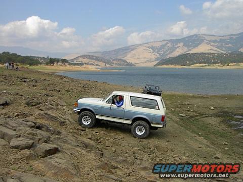 1988 Ford Bronco and another SUV driving on a rocky shore next to a lake