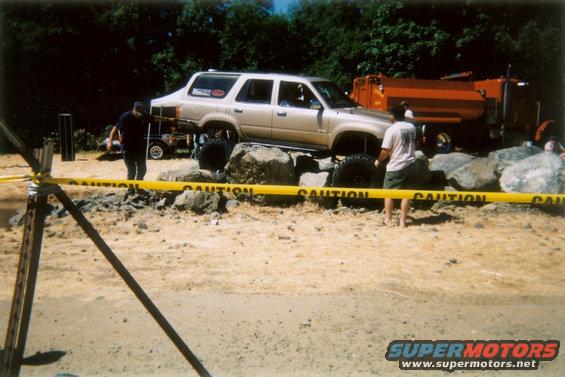 Gold SUV with large tires is on rocks, with a dump truck behind it. Yellow caution tape is in the foreground