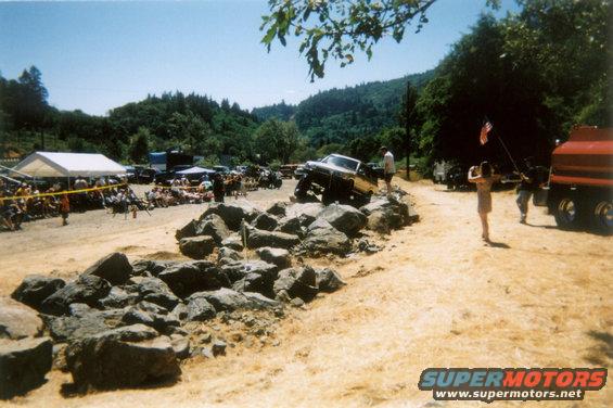 Yellow pickup truck climbing over rocks, 3/4 front view. A crowd watches under a tent