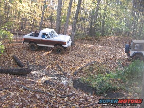 White and brown Ford Bronco II off-roading in woods, another Jeep visible