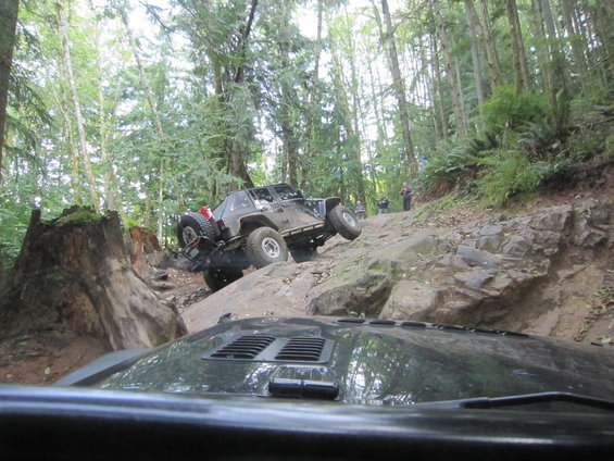 Dark grey Jeep Wrangler climbing a steep, rocky trail in a forest; another vehicle's hood is in the foreground