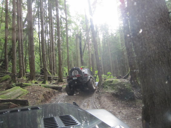 Two Jeeps driving on a muddy forest trail. The front Jeep's hood is visible