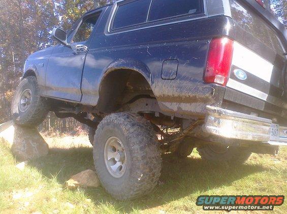 Dark blue 1992 Ford Bronco with off-road tires climbing over rocks, mud splattered on body