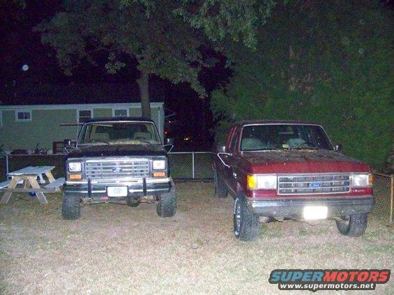 Two Ford Broncos parked in a yard at night. The one on the left is black, and the one on the right is red. Both are facing forward