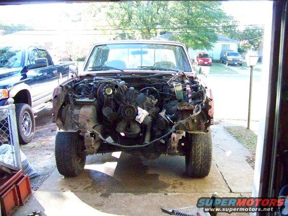 Front view of a disassembled 1980s Ford truck, engine bay empty. Blue Dodge pickup parked to the left