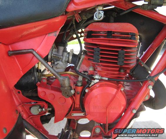 Close-up of a red motorcycle engine with a carburetor and a kick starter. The word HONDA is visible on the engine cover