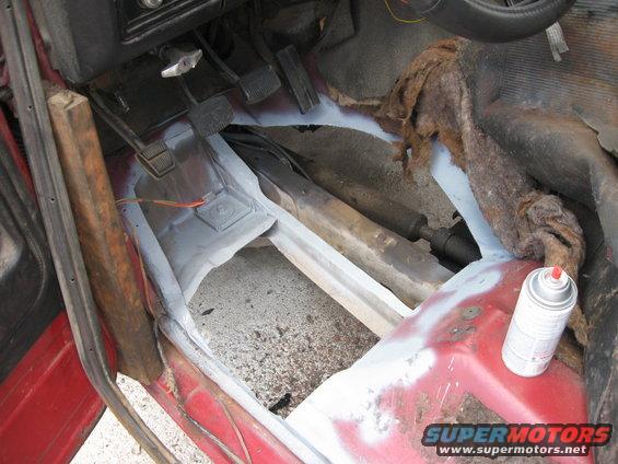 Red vehicle interior floor with rust repair and primer, showing pedals and drive shaft