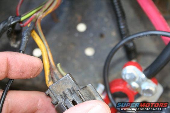 Hand holding a gray electrical connector with multiple wires; battery terminal visible in background