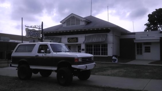 White and brown lifted Ford Bronco parked in front of a building with a 'Drugs' sign