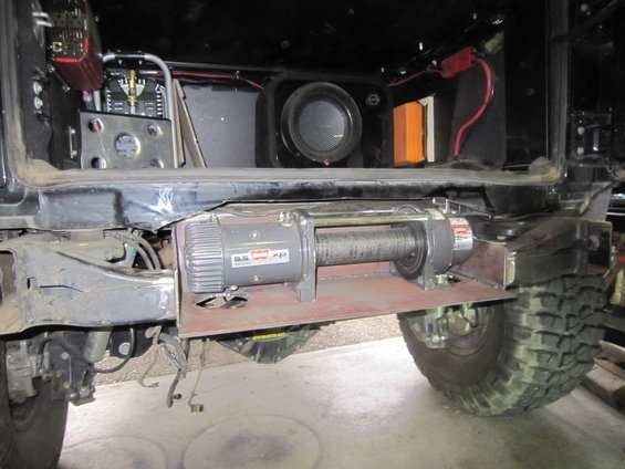 Winch mounted on a metal plate below the rear of a vehicle, with a tire visible