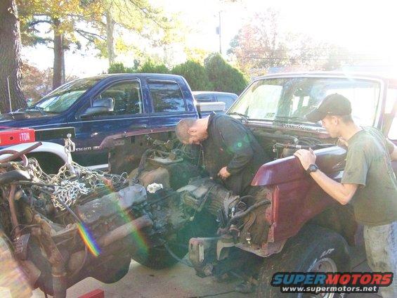 1985 Ford Bronco engine bay with two men working on it. A blue pickup truck is parked nearby