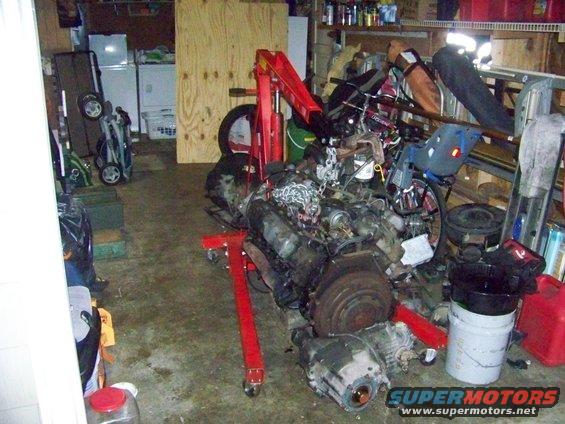 Engine and transmission on a red engine stand in a garage, with laundry machines and shelves in the background