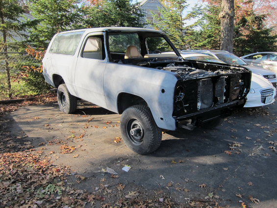 Gray SUV body with hood open, missing grille and headlights, parked on a leaf-covered asphalt lot