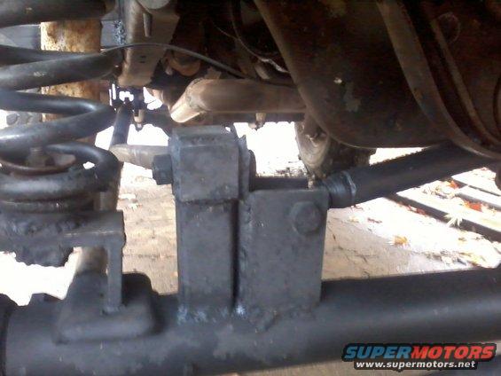 Close-up of vehicle suspension parts, including a coil spring and metal brackets. SUPERMOTORS.NET logo visible