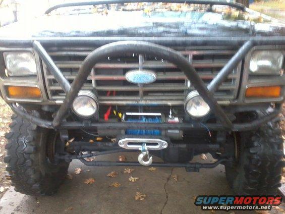 Front view of a modified Ford Bronco with a brush guard, winch, and auxiliary lights. The grille and headlights suggest an early 1980s model