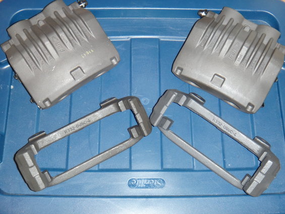 Two gray brake calipers and two gray caliper mounting brackets are laid out on a blue plastic storage bin. Text on brackets reads R332-085C-4
