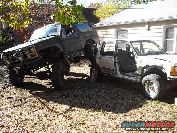 Black Bronco on top of white truck, both heavily modified for off-roading