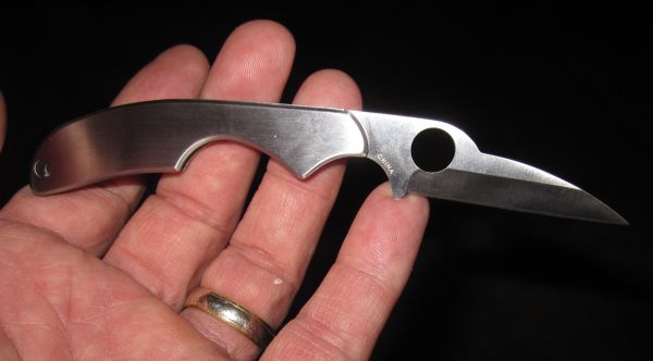 Silver folding knife with a bird-head shaped blade and a circular cutout, held in a hand. Text 'CHINA' is visible