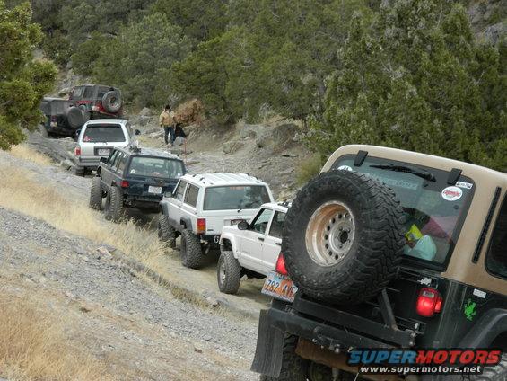 Several off-road vehicles, including Jeeps and SUVs, driving in a line on a rocky, dirt trail with trees and hills in the background. One vehicle has a spare tire mounted on the back
