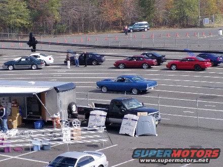 Cars lined up on a track with a trailer and a pickup truck. Some people are standing around