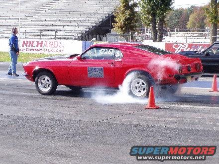 Red 1969 Ford Mustang doing a burnout on a drag strip, rear view