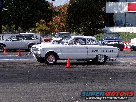 White classic drag car with 'BUMBLE BEE BUILDERS' text on the side, parked on a drag strip with other vehicles in the background