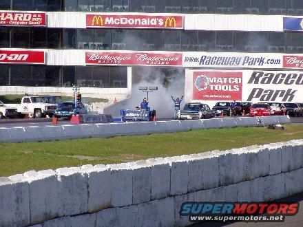 Drag strip with cars lined up, smoke from a dragster, and grandstands with advertisements