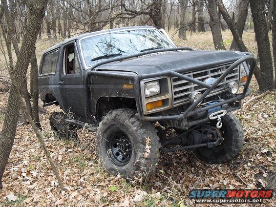 Black modified Ford Bronco with a roll cage and winch, angled from the front-right in a wooded area