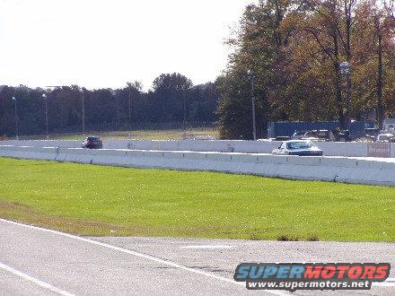 Two cars driving on a racetrack with a grassy area and trees in the background
