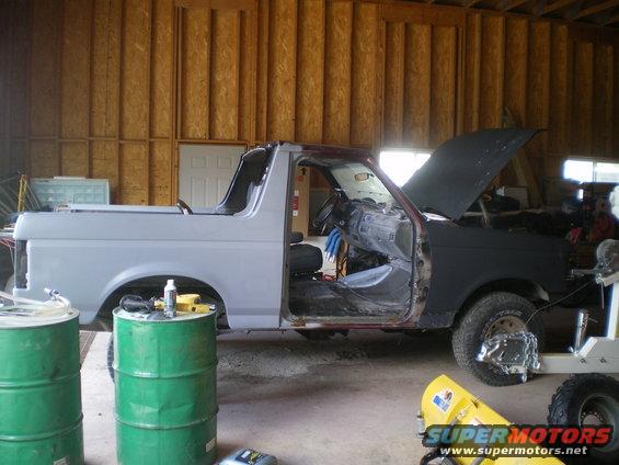 1988 Ford Bronco in garage, doors and hood open, undergoing bodywork. Green barrels and yellow equipment nearby